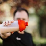 Focus on a man holding a plastic protein shake tumbler outdoors with a blurred background.