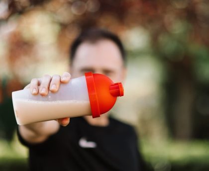 Focus on a man holding a plastic protein shake tumbler outdoors with a blurred background.