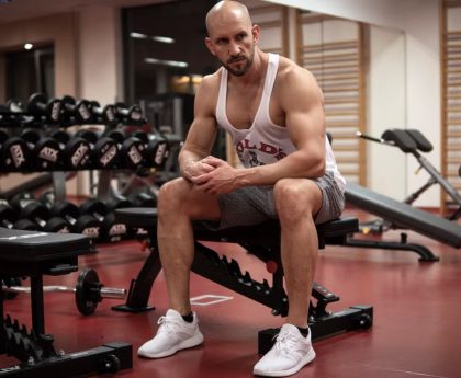 Strong, determined man in white tank top sitting in gym, showcasing fitness and strength.